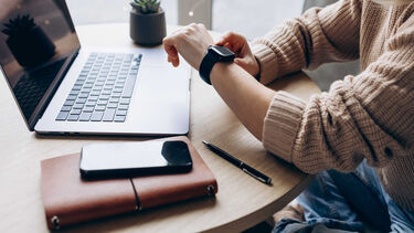A person sits at a laptop and looks at their smartwatch