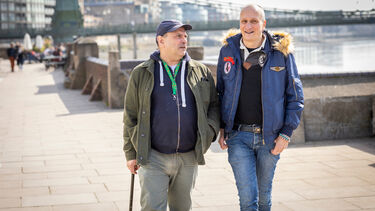 Two men, one with walking stick in front of a river bridge
