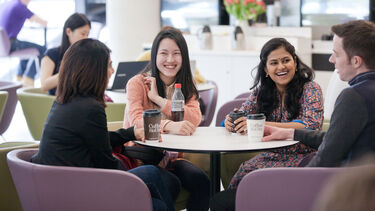Four students sit together at a table in a campus cafe, drinking hot drinks and laughing.