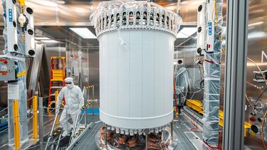 A researcher in protective gear works near a tall, cylindrical scientific instrument with visible wiring. The walls surrounding it are reflective Teflon.