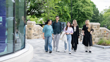 A group of five students are walking towards the camera as they head towards a University building. They are all talking with one another.
