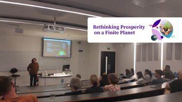 An audience sitting in a lecture theatre while a speaker presents at the front. Overlaid in the top right hand corner is is a circular illustration featuring a close up of a person's hands holdinga small planet. Next to this is the text "Rethinking Prosperity on a Finite Planet"