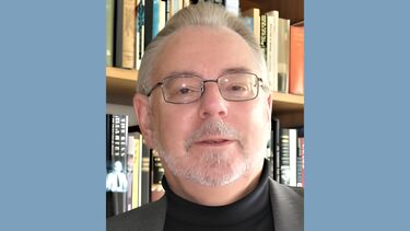 A man with grey hair and glasses stands in front of a booksheld