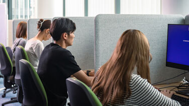 Four students sit in a row in individual computer booths. They are facing away from the camera.