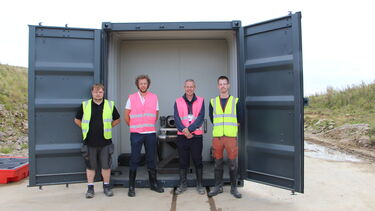 Four men in high visibility vests stand in front of a storage container smiling
