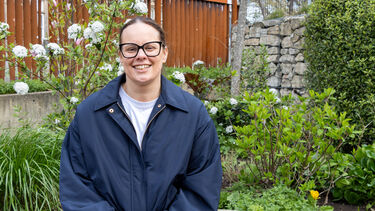 Lisa wears glasses and a blue coat. She has dark hair tied back and sits on a bench in a park.