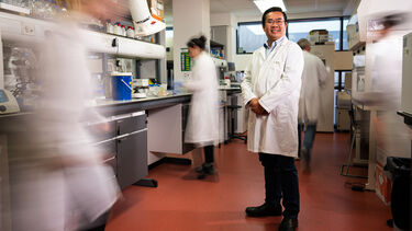 Professor Tuck Seng Wong stands in his lab with his colleagues moving around him in blurred motion