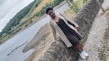 Kemisola wears a black dress and beige cardigan. She leans against a stone wall, and a reservoir is in the background.