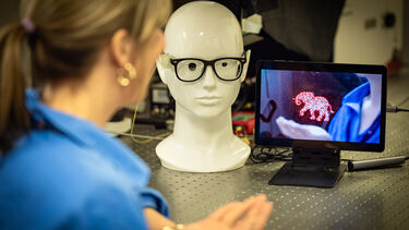 A mannikin head wearing glasses that have been fitted with metamaterials so the wearer could see AR images. There is a tablet at the side of the mannikin head displaying what the wearer would see - this is showing a woman's hand holding a hologram of a unicorn.