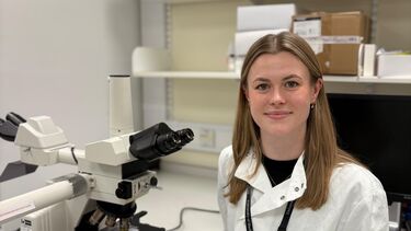 A person smiling in a lab coat sitting by a microscope