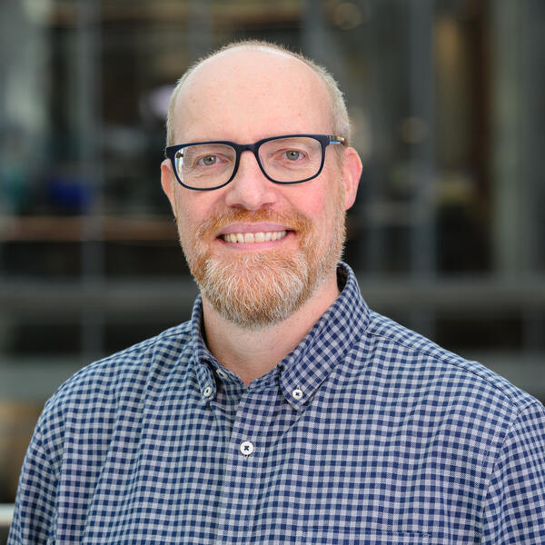 Profile picture of Profile of an academic wearing a blue checked shirt and black glasses