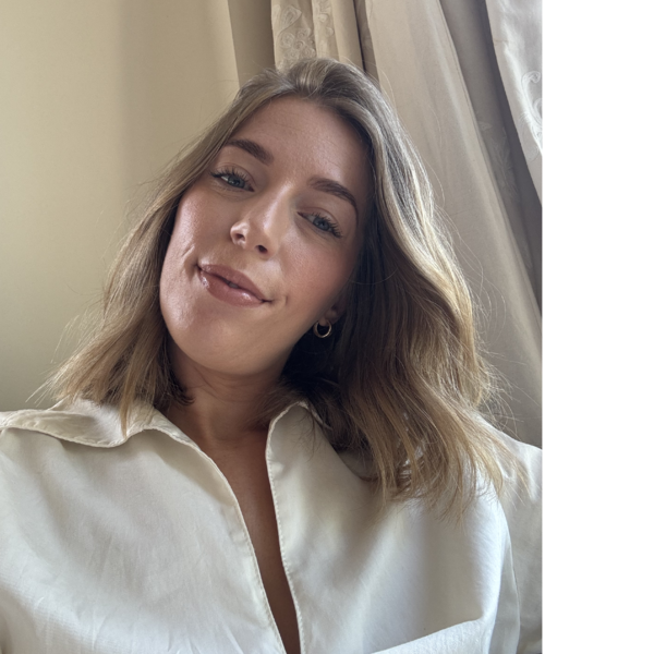 Profile picture of Kate looks down at the camera, wearing a white shirt, with a curtain in the background