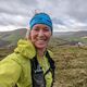 A woman wears a blue headband, a running backpack and a lime green jacket. She holds the camera for a selfie, while behind her are rolling hills and a sheep.