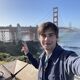 A male student stands outside with the Golden Gate Bridge in the background. He is smiling at the camera and making a peace sign with his hand.