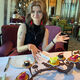 A female sits at a table in a restaurant and there is table of small cakes in front of her. She holds her hands up to gesture towards the cakes and smiles at the camera.