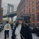 A female student stands in the foreground of an urban street in the USA, wearing a baseball cap and smiling at the camera. Behind them is a bridge in the background.