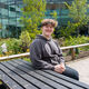 Alfie wears a grey hoodie and sits at a picnic bench. The SU and lots of plants are behind him.