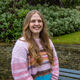 Charlotte has long blonde hair. She wears a pink stripy jumper and stands in front of some trees and bushes.