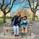 A sunny autumnal/wintery day; a woman wrapped up in a black puffer jacket, scarf, gloves and light jeans sits smiling on a bench in a park leaning in towards a Paddington Bear statue. The statue is eating the classic marmalade sandwich, which the character is known for. Paddington is wearing his classic blue toggle coat, and red hat. Behind them on the bench, there are wintry trees missing their leaves and a few obscure buildings.