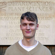 Image of Dr. Christopher Bousfield in front of a beige stone sign reading department of plant sciences