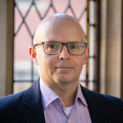 A person without hair wears glasses. They're in front of leaded windows