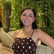 A female stands with her hand on her hip and smiles towards the camera. Behind her are some trees.