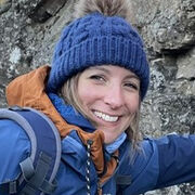 Helen wears a blue bobble hat and blue jacket, and she stands in front of a stone wall.