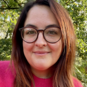 Emma faces the camera in front of a tree, wearing glasses and a pink jumper