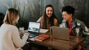 Three people laughing while sitting at a table with two laptops