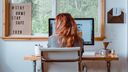 Woman working from home, sitting on a white chair in front of a computer.