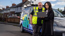 a man in a hi-vis vest looks over the shoulder of a woman, a researcher, who is holding a laptop. In the background is an electric van. 