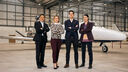 four people, one asian man, a white woman, a white man and an asian woman, stand in a hanger with a small plane in the background