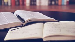 An open printed book, a pen and an open notebook on a table. In the background is a bookshelf.