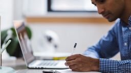 A photo of a man at a desk, writing into a notebook. Beside him is a laptop.