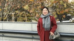 Alumni Xin Feng standing in front of a tree and a building