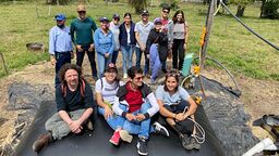 a group of people sit in a field, surrounded by trees. They sit on a black square of tarpaulin and are next to some other equiment like pipes and hoses. 
