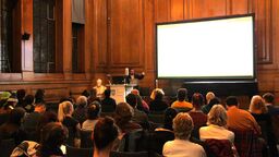 Gee at the front of the room with a PowerPoint slide on a projector. The audience is sat in front of the projector. 