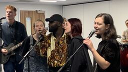 Five students singing behind 3 microphones. One plays a guitar