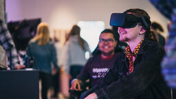 A man wearing a VR headset looks into space just off camera, he is smiling and enjoying the activity.