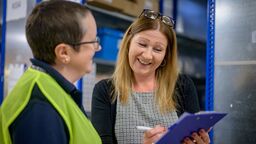 Two women holding a clipboard in a warehouse, one is wearing a high vis vest