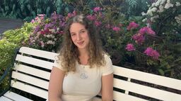 A female student sits on a white bench and smiles at the camera. Behind her are trees with pink and white flowers on them.