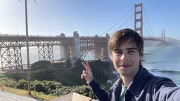 A male student stands outside with the Golden Gate Bridge in the background. He is smiling at the camera and making a peace sign with his hand.