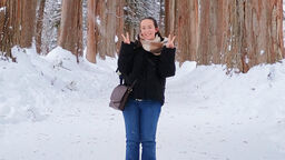 A female student stands on a snowy path with trees lined either side of the path and behind, branches filled with snow. She smiles at the camera and makes a peace sign with both hands.