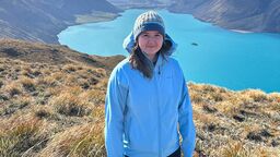 A female student wearing a blue winter hat and blue waterproof jacket stands smiling at the camera on a hill. In the background is a large lake with mountainous backdrop.