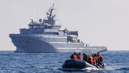 A small boat crossing the sea, in the background a much larger military or security type vessel can be seen.