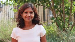 A person smiling at the camera outside, surrounded by trees and bushes.