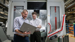 Two men stand in front of a manufacturing machining centre looking into a laptop.
