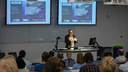 A man wearing a dark grey suit, dark framed glasses and a rainbow lanyard is stood at a lectern speaking to lecture theatre full of people. Behind him are two projector screens.