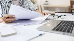 A person reviewing papers at a desk, with a pair of glasses and a laptop nearby