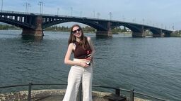 A female student stands in front of a body of water and smiles at the camera whilst holding a drink in a bottle. There is a bridge in the background.